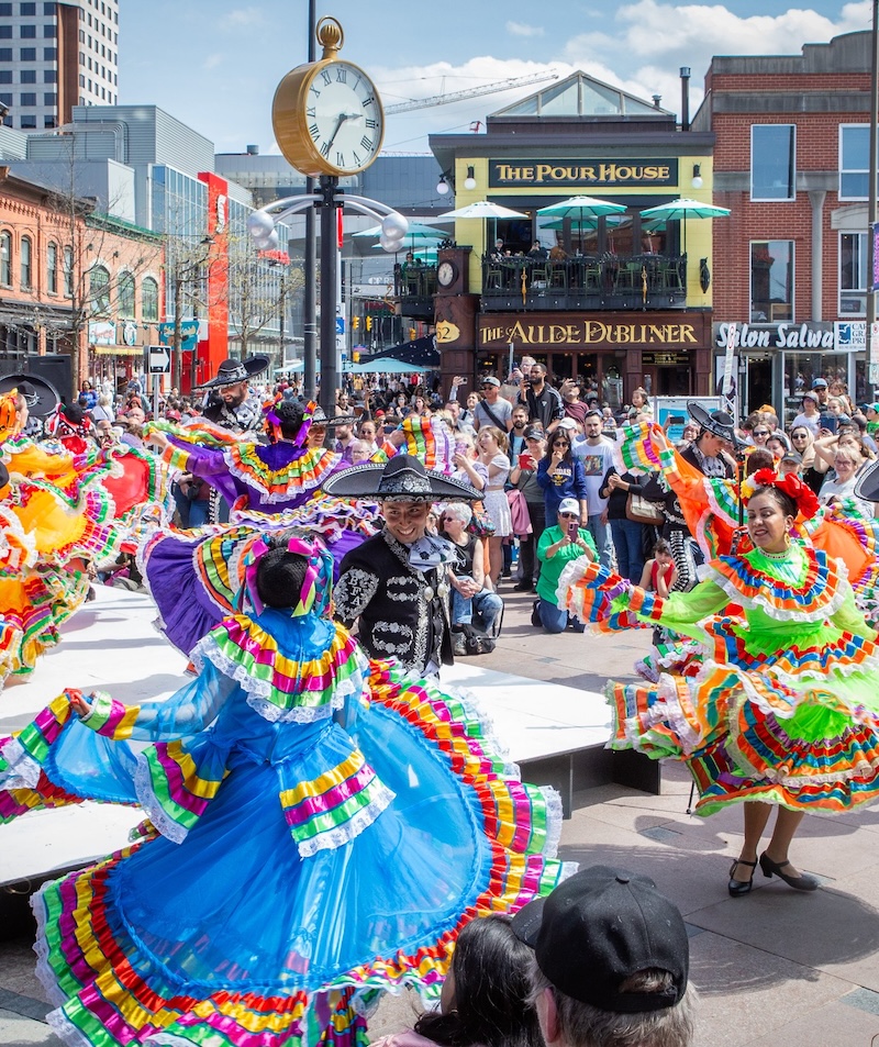 Des danseuses vêtues de costumes traditionnels aux couleurs vives se produisent lors d'un festival en plein air très animé. La foule les regarde, avec en arrière-plan des bâtiments historiques en briques, des restaurants et une grande horloge de rue, par une journée ensoleillée.