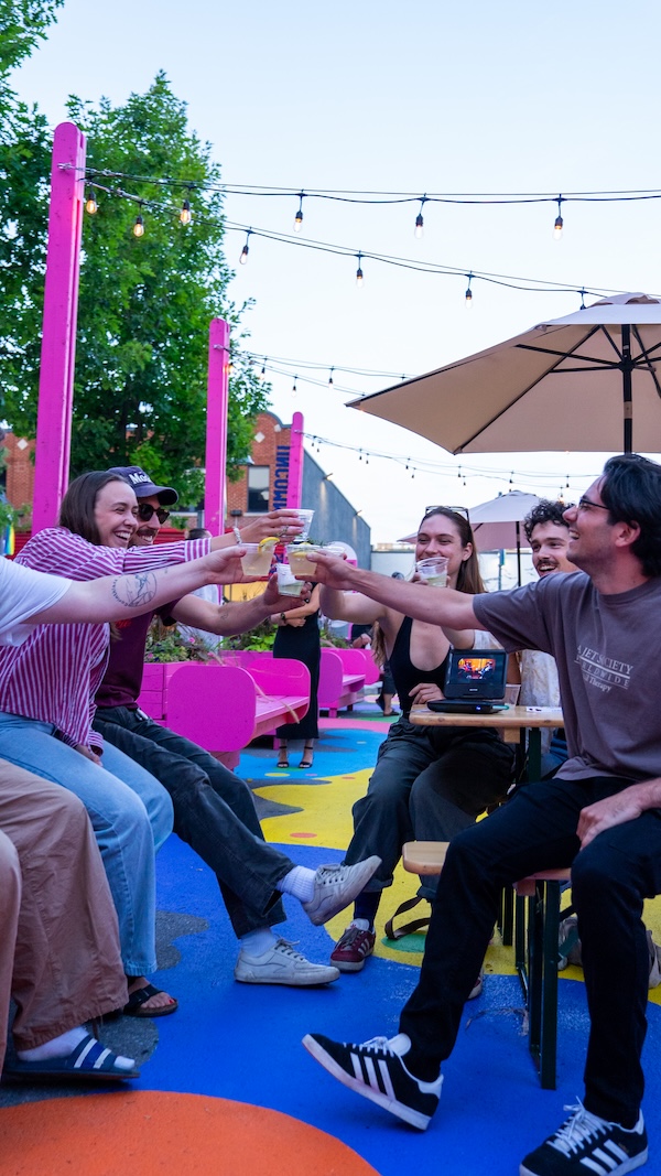 Un groupe d'amis, assis à l'extérieur autour de tables colorées, trinquent en souriant et font la fête sous des guirlandes lumineuses, sur une terrasse lumineuse et animée.