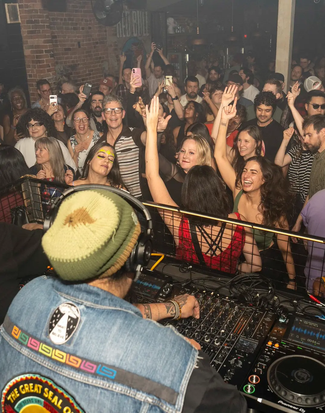 Dans une boîte de nuit, une foule animée danse et sourit tandis qu'un DJ coiffé d'un bonnet mixe sur ses platines ; certaines personnes ont les mains en l'air et l'ambiance est dynamique et festive.